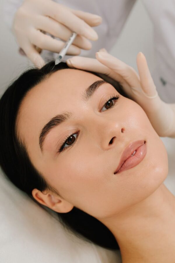 Eine Frau liegt auf einer Behandlungsliege, während eine medizinische Fachkraft mit Handschuhen eine Injektion in die Kopfhaut durchführt.